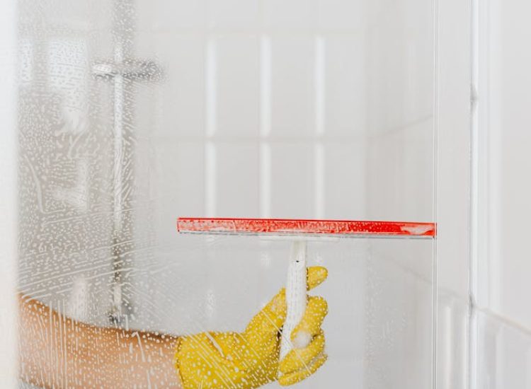 A person wearing a yellow glove cleans a shower glass with a squeegee, emphasizing hygiene and cleanliness.